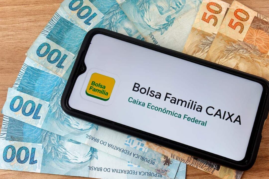 Imagem de um celular aberto com o aplicativo do Bolsa Família em cima de uma pilha de dinheiro.