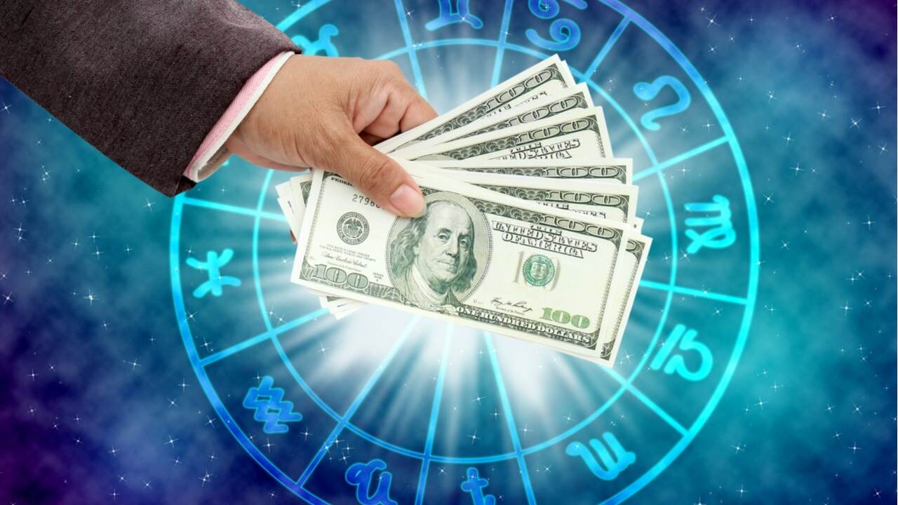 Na imagem, mão segurando dinheiro em frente aos símbolos de cada signo do horóscopo