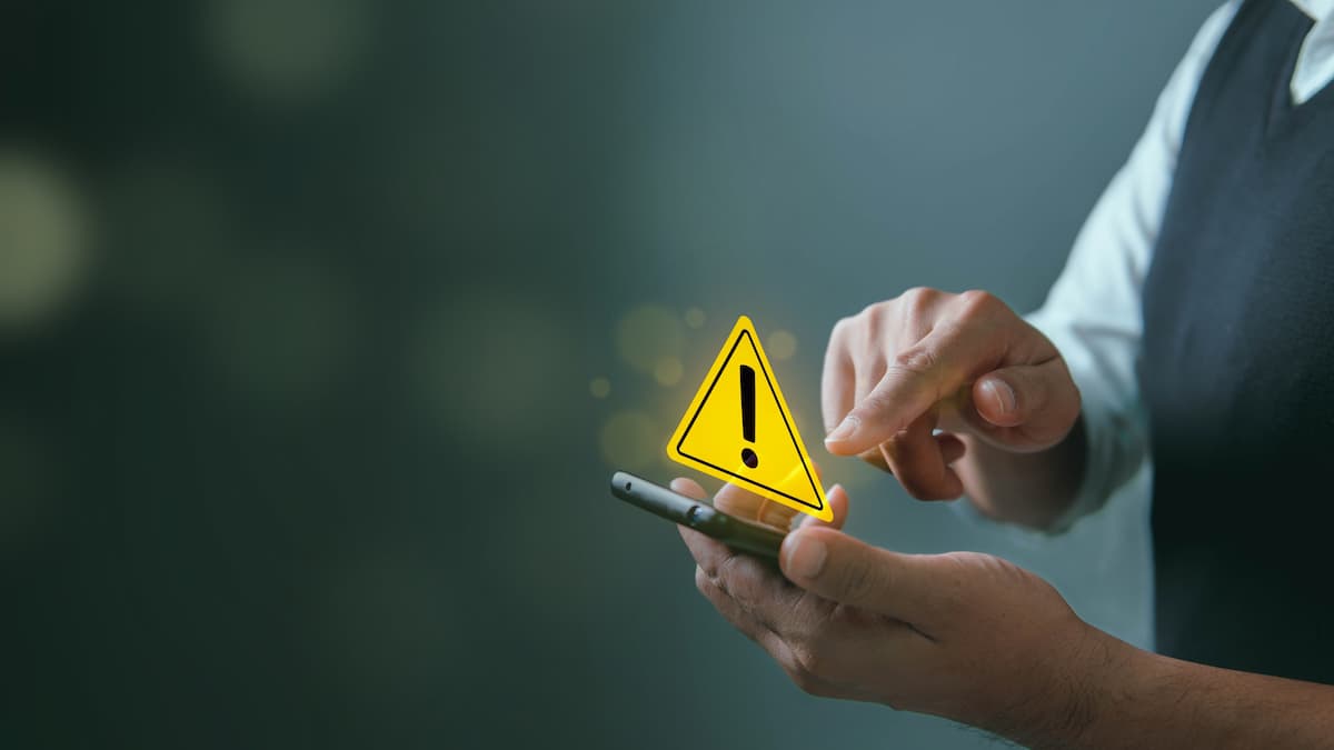 Mãos segurando e mexendo em celular. Em cima da tela do celular, como se fosse um holograma, há o símbolo amarelo de alerta, com uma exclamação