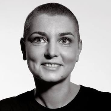 Imagem de Sinéad O'Connor, cantora famosa mundialmente que faleceu. A foto está em preto e branco enquanto ela sorri, com o cabelo raspado