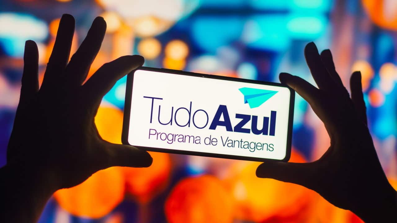 Duas mãos segurando celular que mostra logo do programa de vantagens e pontos TudoAzul. Ao fundo, luzes desfocadas.