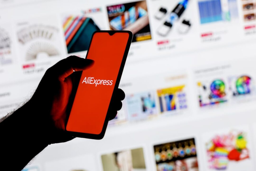 Mão segurando celular com logo do AliExpress na tela e, ao fundo, um site com produtos a venda