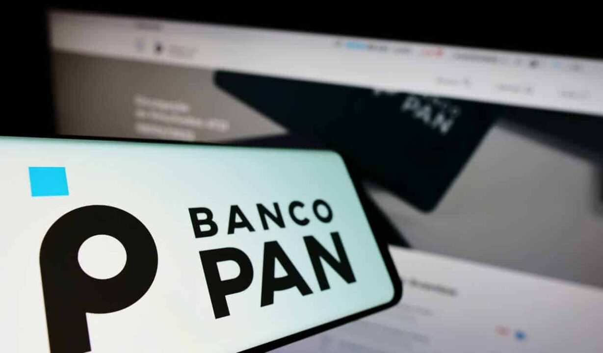 Celular com a logo do Banco Pan na tela, ao fundo um notebook com o site do mesmo banco aberto