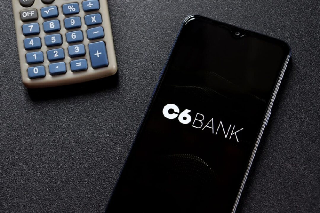 Imagem de um celular com a tela na logo do C6 Bank. Ao lado, uma calculadora.