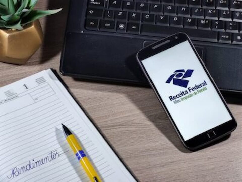 Imagem de um celular com a tela na logo da Receita Federal Imposto de Renda e um caderno ao lado.