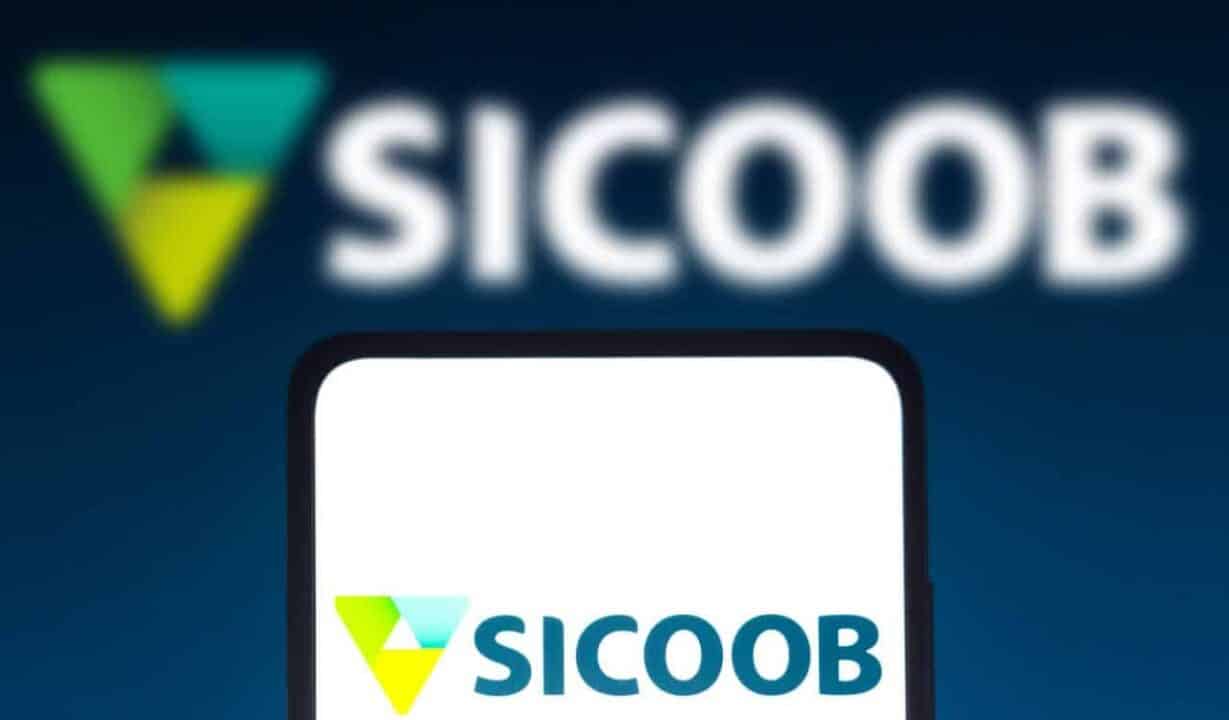 Celular com do Sicoob na tela. No topo da imagem, em segundo plano, aparece a logo da cooperativa desfocado