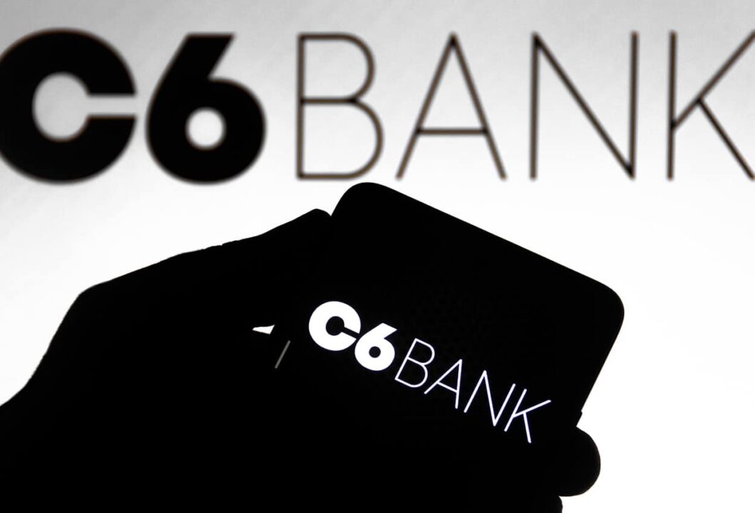 Silhueta de uma mão segurando um celular que exibe o logotipo do C6 Bank. No fundo da imagem, aparece o nome do banco digital.