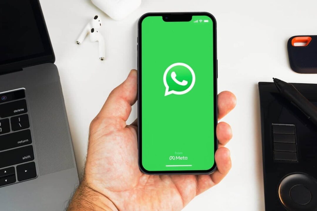 Mão segurando celular com app WhatsApp aberto. Ao redor, fones de ouvido, caneta e notebook.