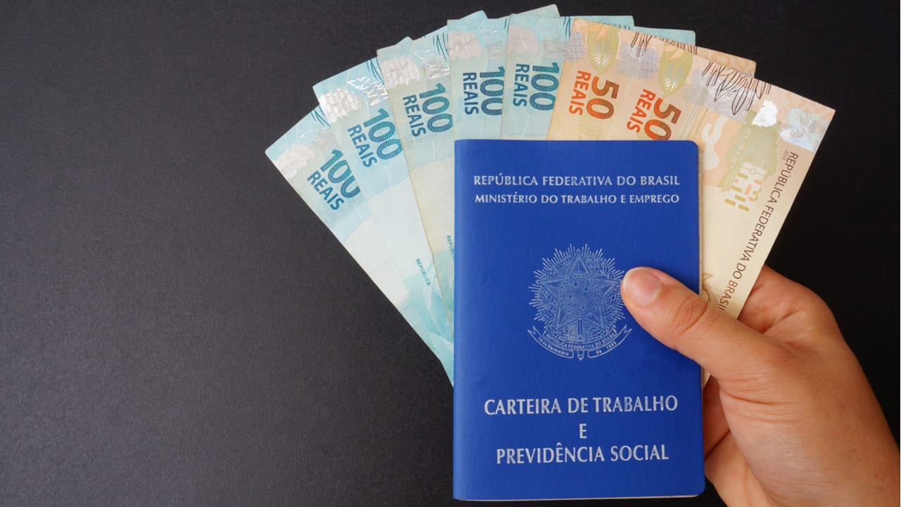 Imagem de mão segurando uma carteira de trabalho, com notas de 50 e 100 reais dentro dela, representando reajuste do salário mínimo