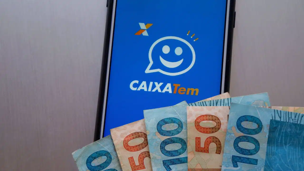 Notas de 50 e 100 reais sobre um celular com app Caixa Tem aberto.