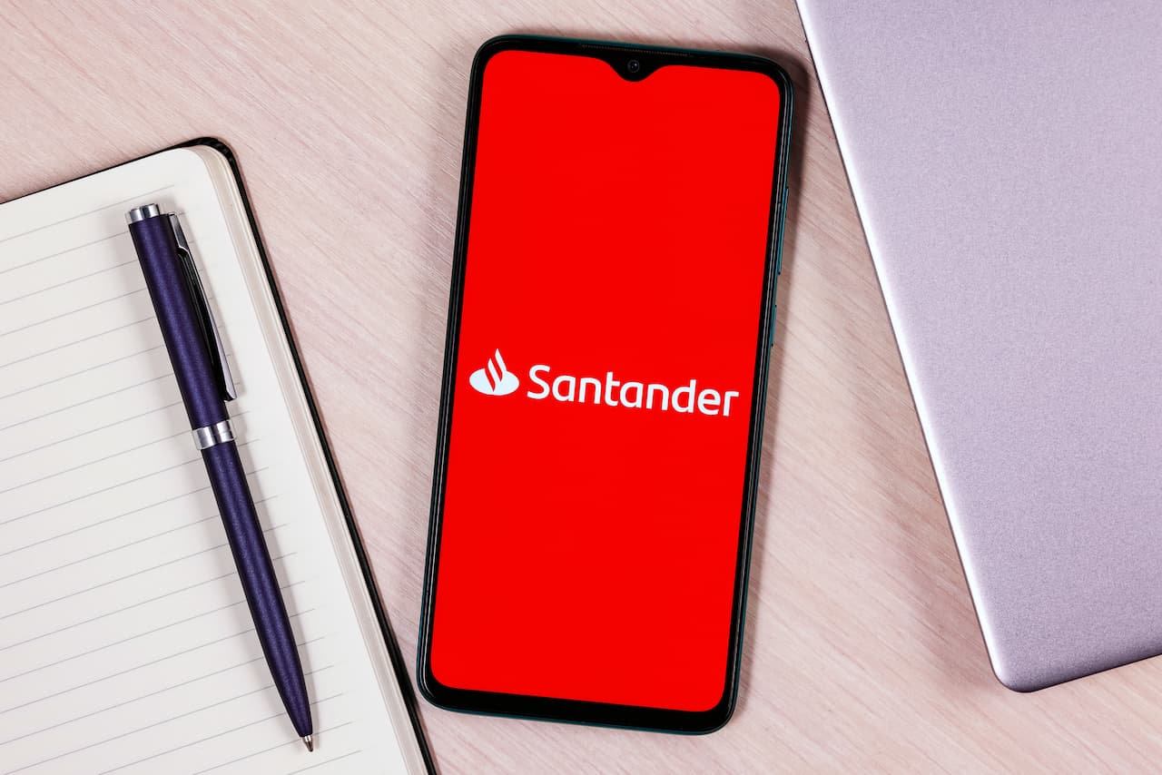 Celular com logo do Santander na tela ao lado de laptop, caderno e caneta