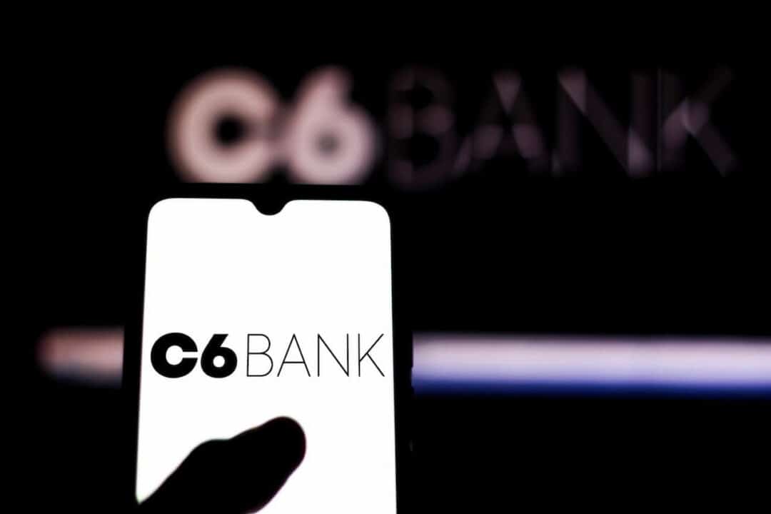 Mão segurando celular com aplicativo do C6 Bank aberto e, ao fundo, logo do C6 Bank em um telão.