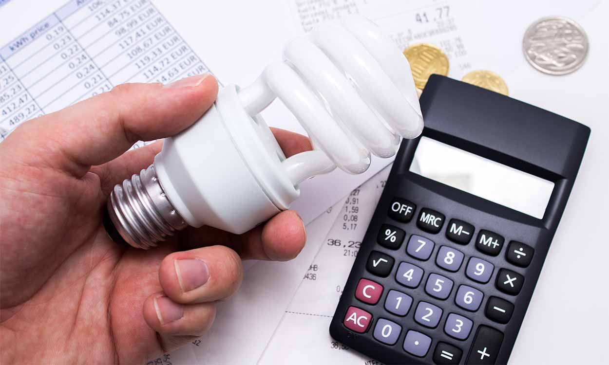Mão segurando uma lâmpada. Abaixo, em uma superfície, uma conta de energia, outros papéis, uma calculadora e moedas, representando o pagamento da conta de luz