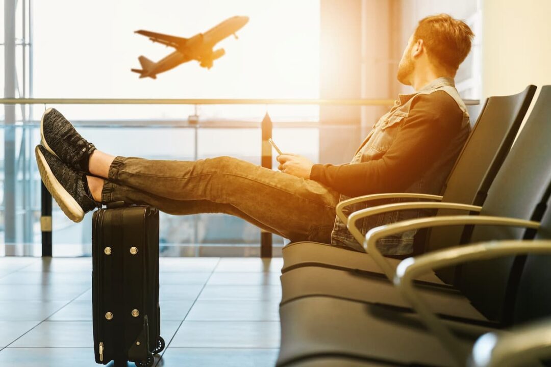 A imagem mostra um homem sentado no aeroporto, esperando o voo. Vendo pela janela um avião decolar.