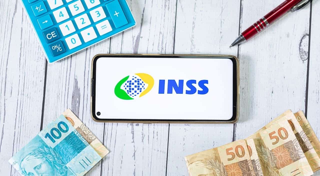 Celular com logo INSS e calculadora, caneta e cédulas brasileiras ao redor, representando auxílio de R$ 1.320