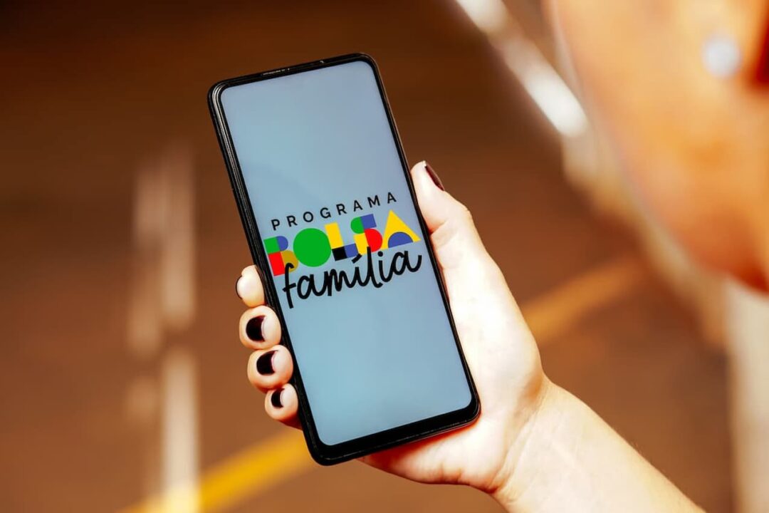 Mão com esmalte preto segurando celular com logo do Bolsa Família na tela.