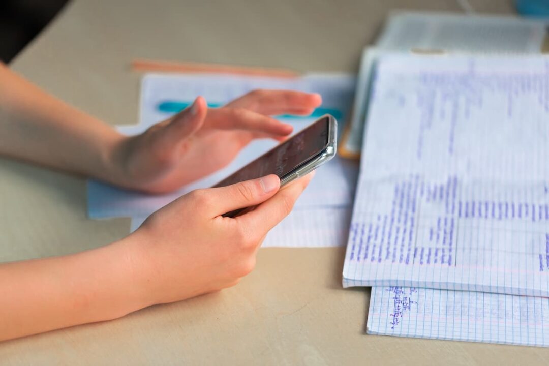 Estudante mexendo no celular em uma mesa escolar com material de estudo