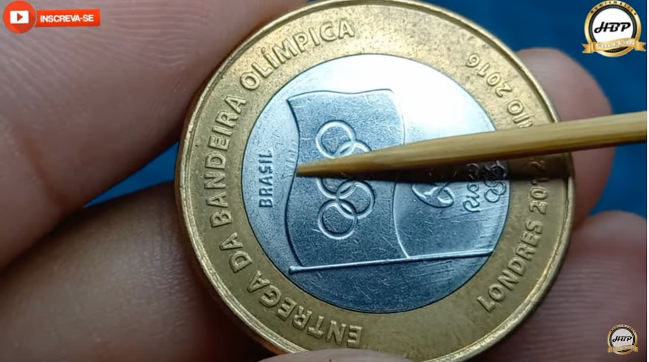 Mão segurando uma moeda de R$ 1 com um palito indicando o erro de fabricação