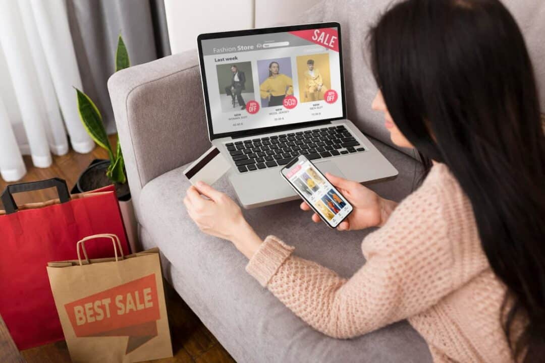 A imagem mostra uma mulher com o notebook aberto em um site com ofertas e o celular também. Além de ter sacolas de comprar ao seu redor e segurar um cartão de crédito para finalizar compra.