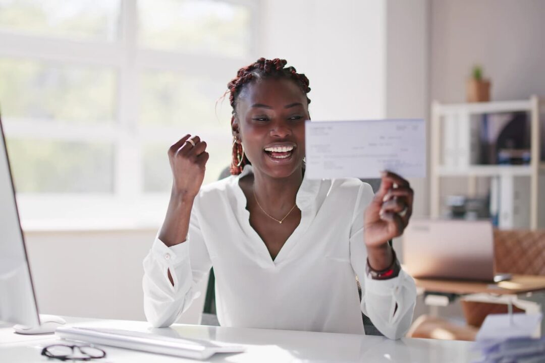 mulher negra com tranças sentada em uma escrivaninha muito feliz com um cheque na mão