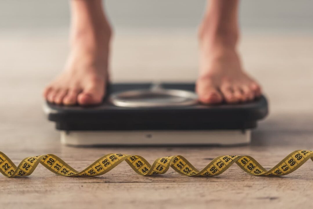Fita métrica em primeiro plano. Ao fundo, pés humanos sobre uma balança