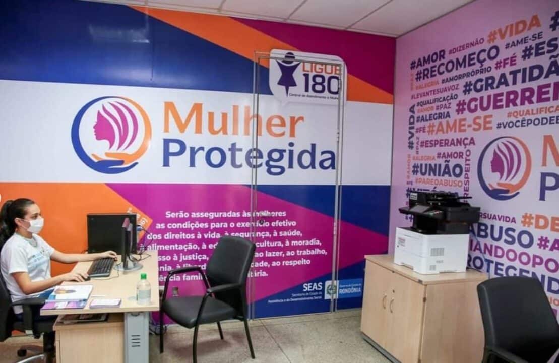 Mulher em uma sala, cuja parede tem a logo e o nome do programa do estado de Rondônia "Mulher Protegida".