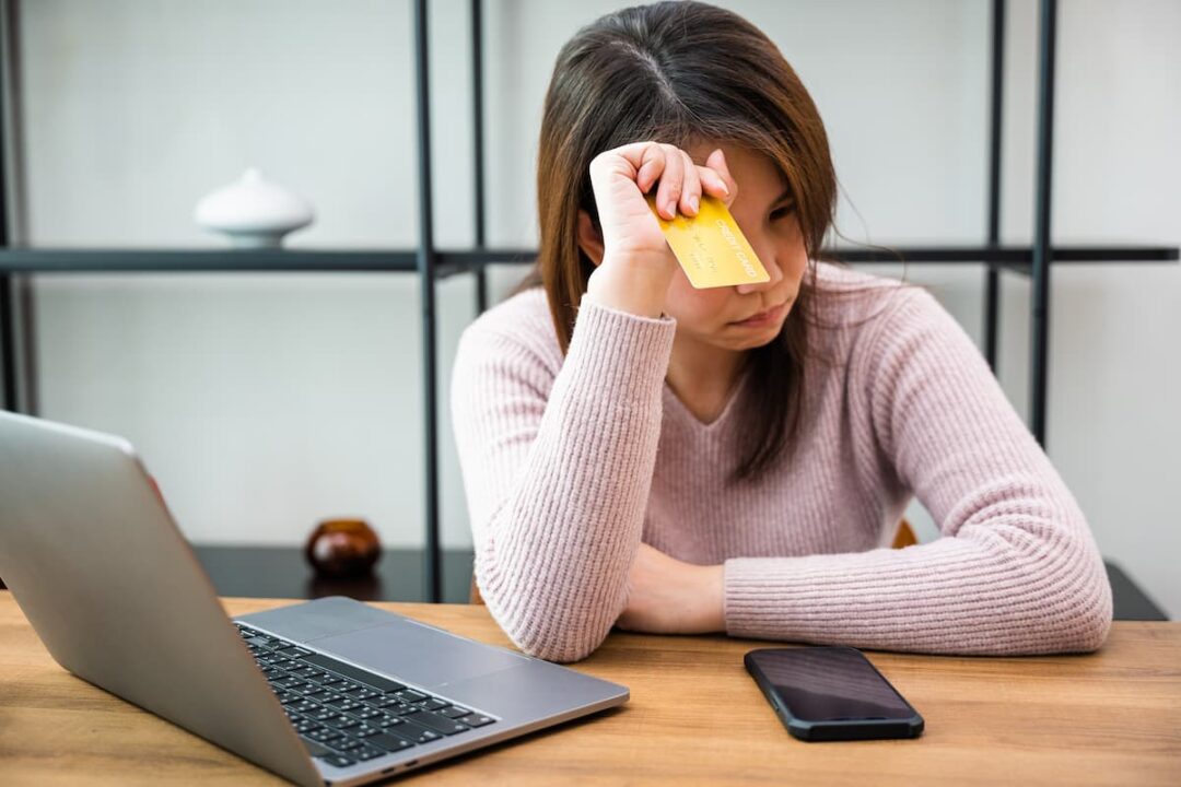 Cliente de banco insatisfeita com cartão de crédito em mãos, apoiada sobre mesa com celular e notebook na sua frente