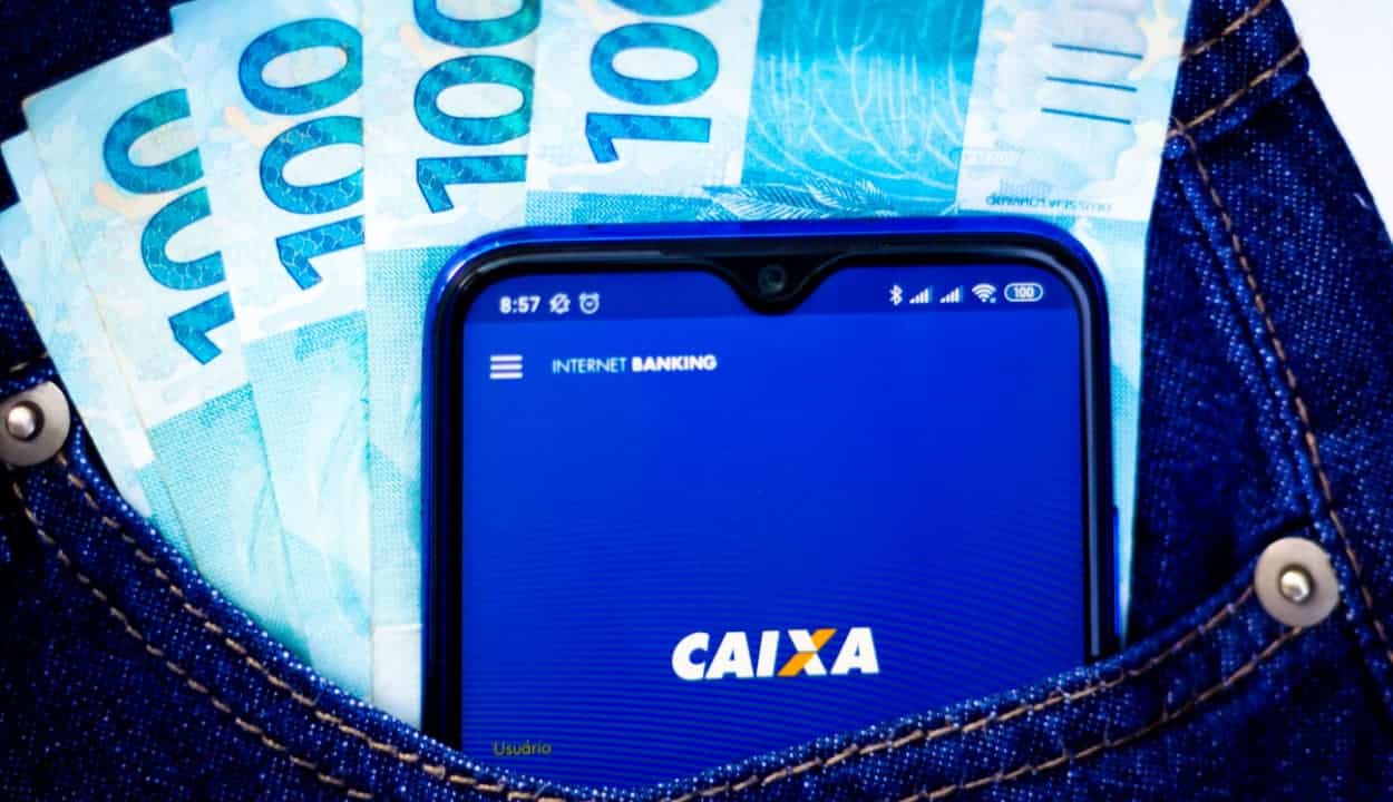 Celular com app da Caixa aberto e notas de 100 reais em um bolsa de uma roupa jeans