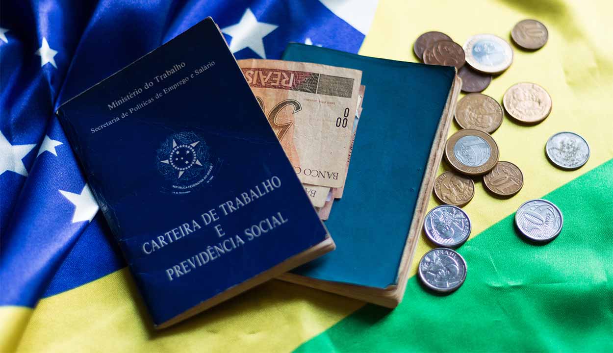 Carteira de trabalho sobre bandeira do Brasil ao lado de moedas e cédulas de real ilustrando seguro-desemprego