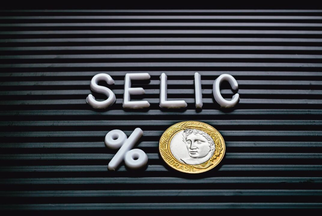 Sobre uma superfície preta, letras brancas formando a palavra Selic. Abaixo, um símbolo de porcentagem e uma moeda de 1 real.
