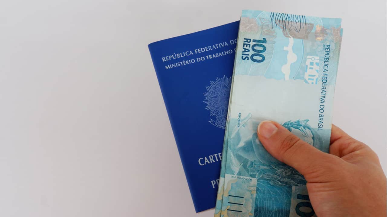 Mão segurando carteira de trabalho e notas de 100 reais
