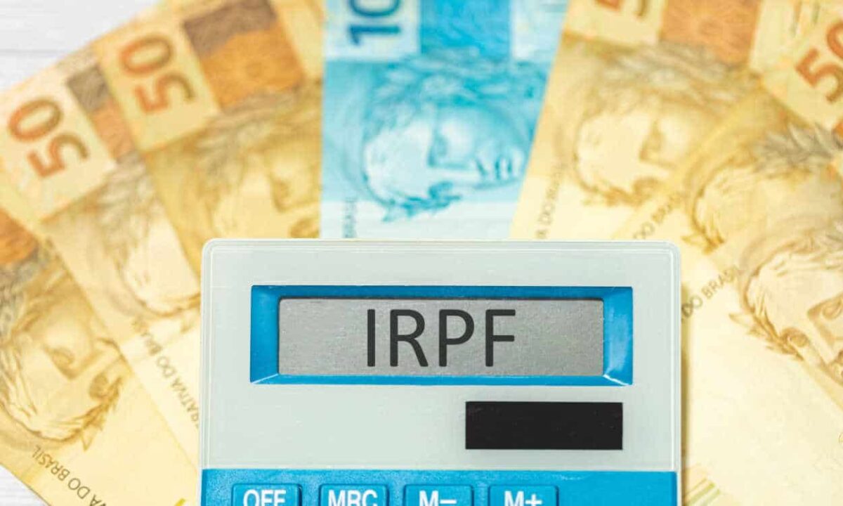 Imagem de uma calculadora branca e azul com a sigla IRPF na tela sobre notas de 50 e 100 reais em formato de leque.