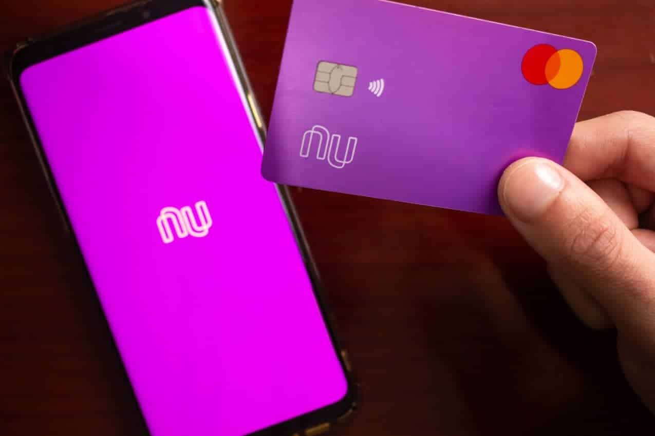 Celular com aplicativo do Nubank na tela inicial e ao lado, mão segurando o cartão da instituição.