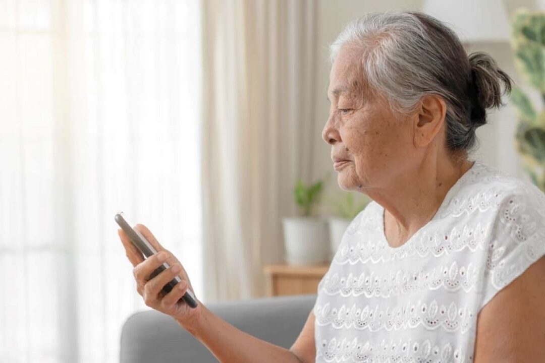 idosa sentada em um sofá, enquanto usa o celular para fazer prova de vida