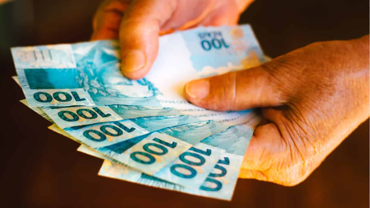 Mão segurando notas de 100 reais de auxílio