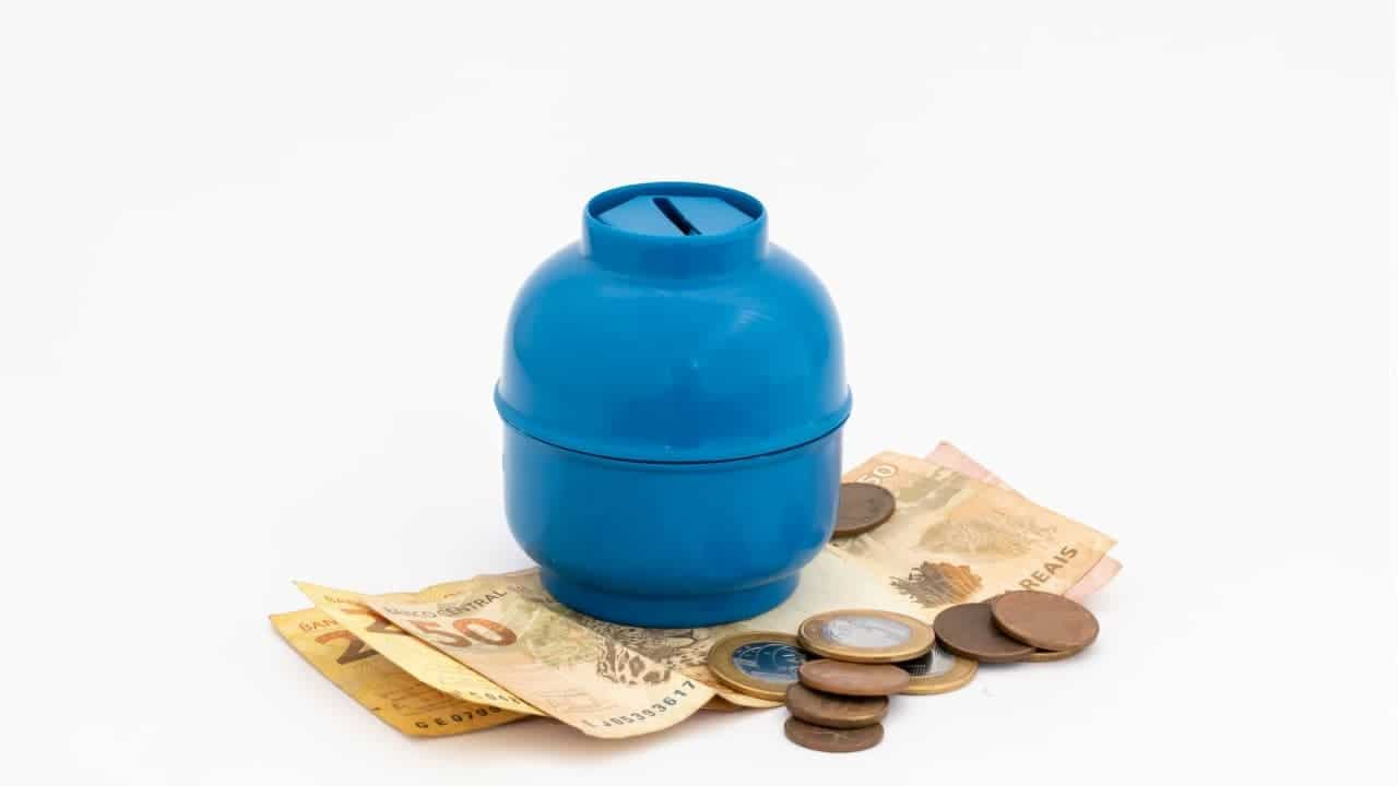 Cofrinho em formato de botijão de gás em cima de notas de 50 e 20 reais e moedas ao lado, que representam auxílio gás auxilio-gas