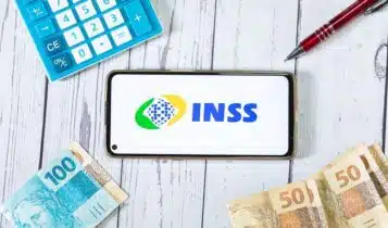 Celular com logo INSS e calculadora, caneta e cédulas brasileiras ao redor