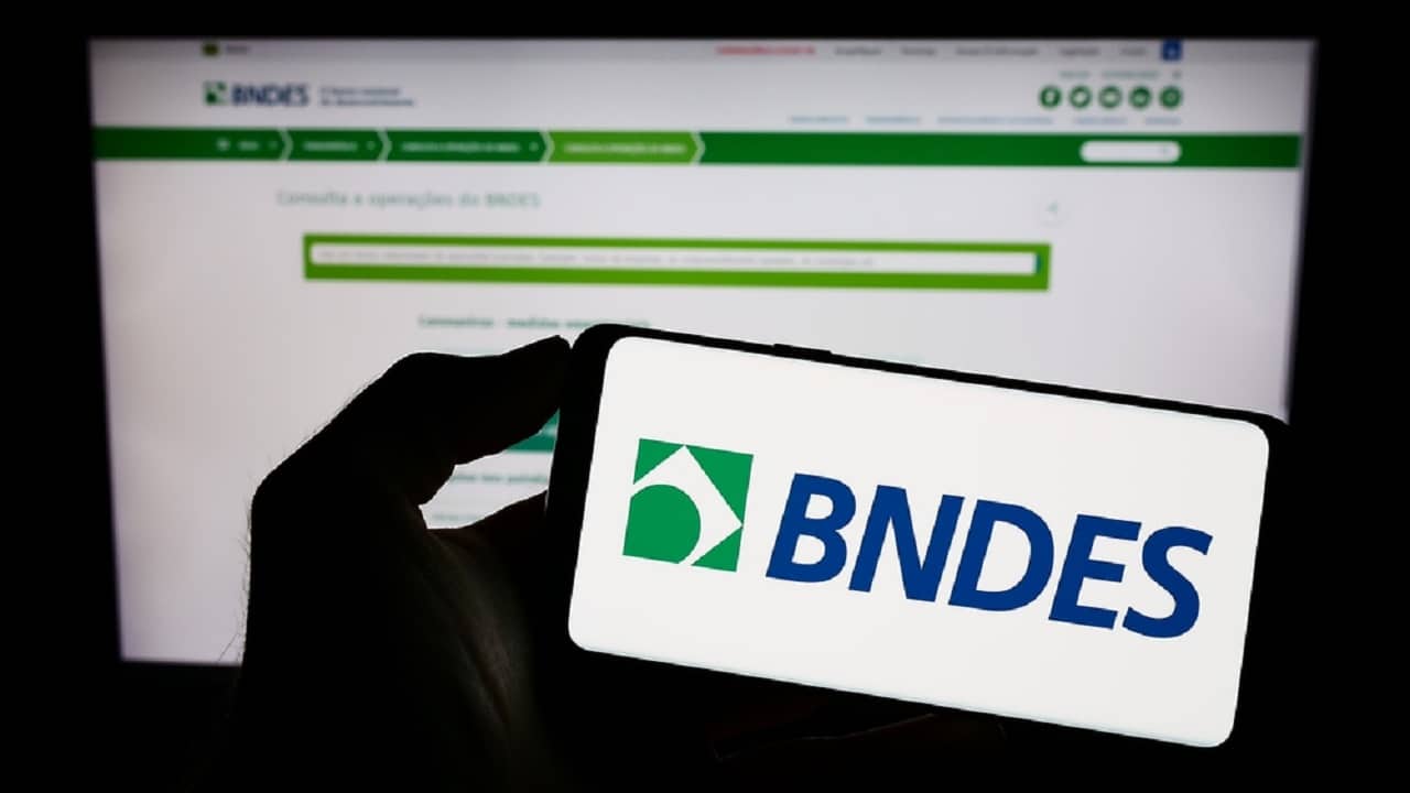 Celular com logo do BNDES e computador no fundo