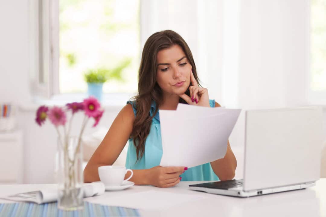 Na imagem, mulher com papéis na mão e um laptop na mesa.
