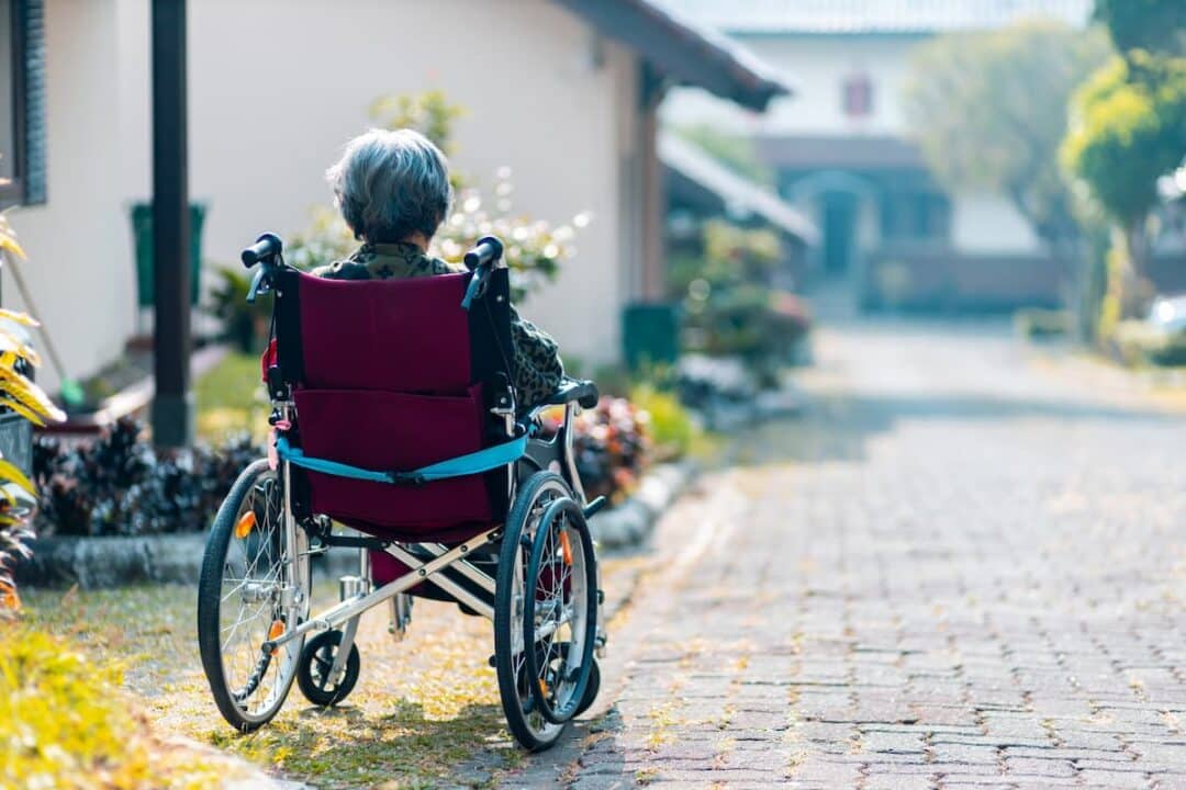 A imagem mostra uma senhora de costas, sentada em uma cadeira de rodas, na rua.