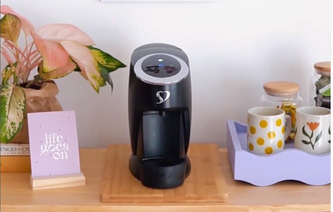 Uma cefeteira preta da marca três corações ao centro. Do lado direito um bandeija contendo canecas e potes; do lado esquerdo uma planta, livros e um quadro.