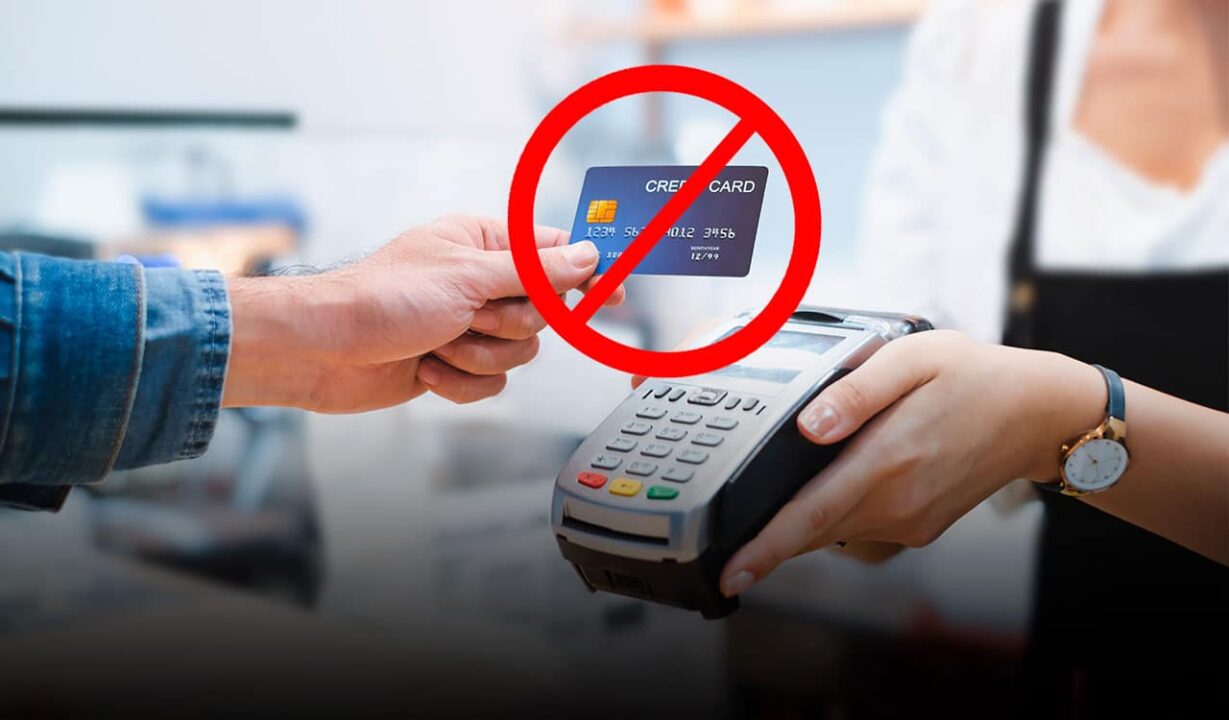 Pessoa utilizando um cartão para pagar algo em uma maquininha, em cima do cartão há um símbolo de 'proibido'.
