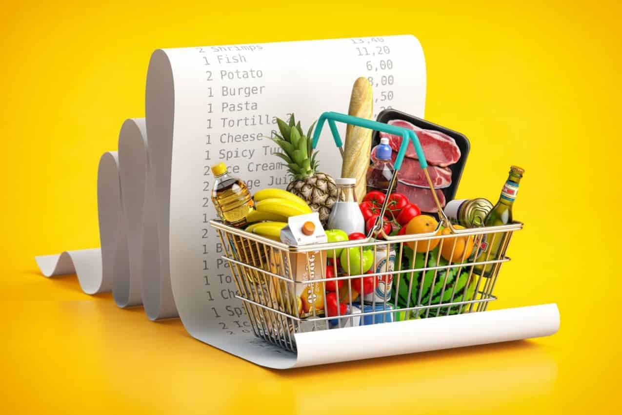 Cesta de compras com alimentos de uma cesta básica em cima de uma nota fiscal enrolada sobre fundo amarelo.