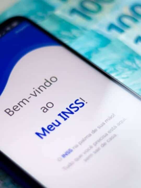 Celular mostrando tela inicial do aplicativo Meu INSS sobre um conjunto de notas de R$ 100
