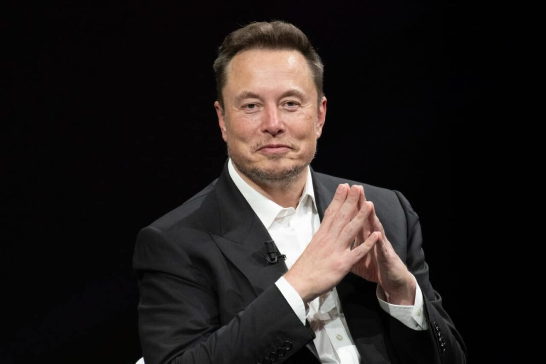 Elon Musk com as mãos juntas sorrindo para a foto, representando o interesse do bilionário em entrar no mercado de telecomunicações.
