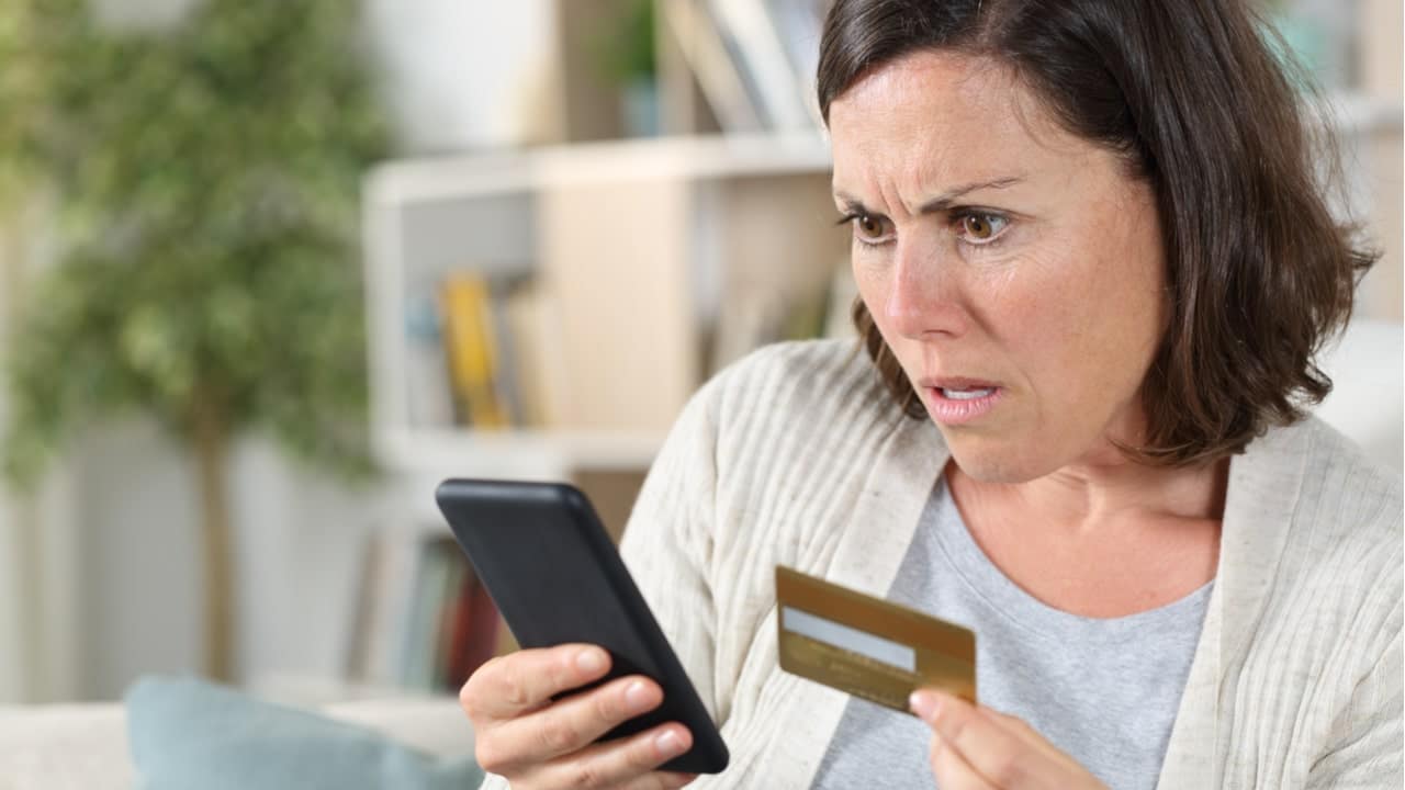 Mulher segurando cartão e celular com expressão de incredulidade no rosto em alusão ao golpe financeiro.