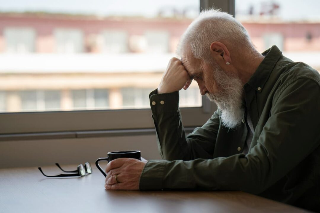 A imagem mostra um homem idosos, com a cabeça baixa, apoiada em uma das mãos, aparentemente triste.Demência