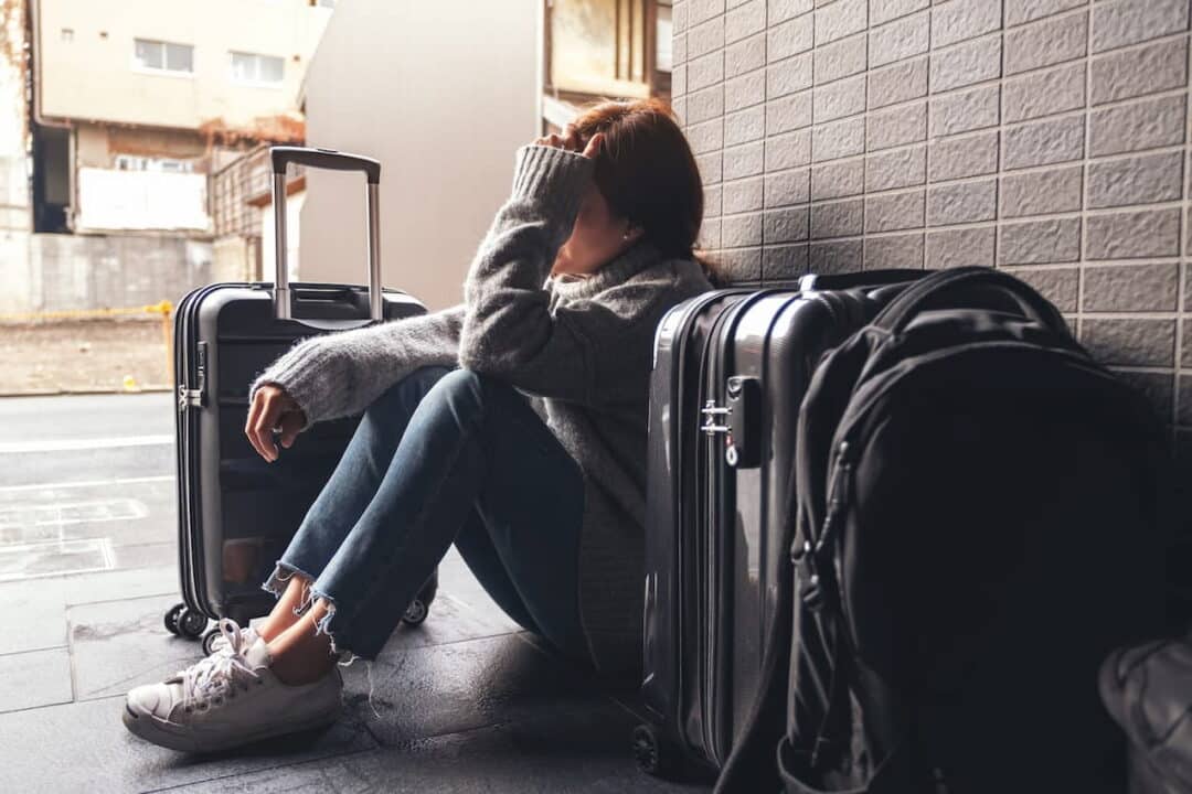 Mulher sentada no chão do aeroporto, com a mão no rosto, preocupada, com malas de viagem ao redor.