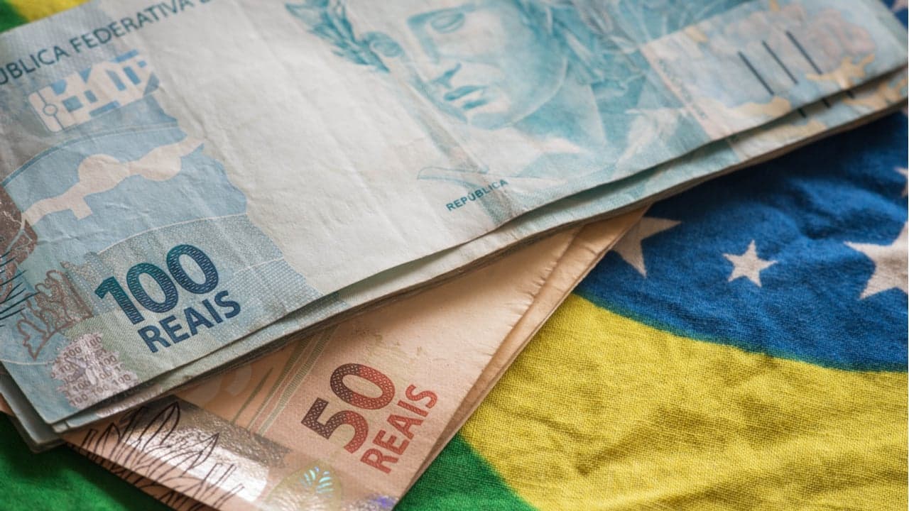 Imagem de notas de 100 e 50 reais, sobre uma bandeira do Brasil.
