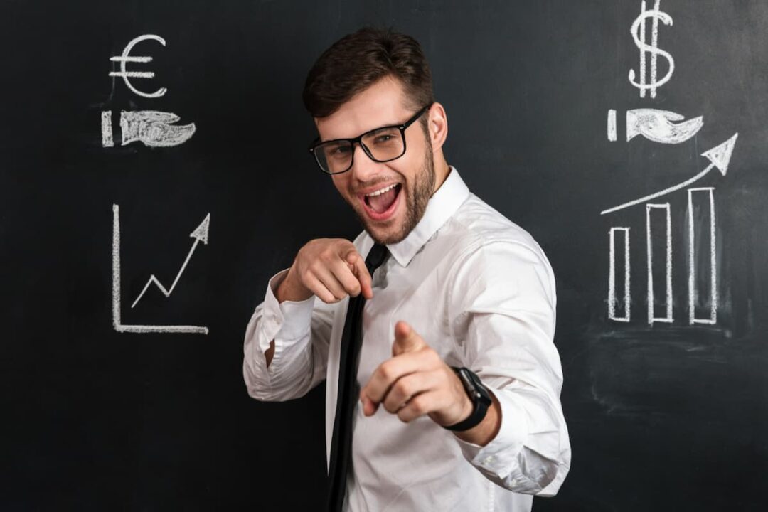 A imagem mostra um homem, com óculos, roupa social, apontando para a frente, com expressão de feliz e confiante. Ao fundo, aparecem dois gráficos indicando subida de valores com o símbolo do Euro e dólar.